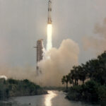 Apollo 13 Blasted Off 55 Years Ago – NASA Anniversary Apollo 13 launches upward, leaving a column of flames and light brown vapor behind it. Trees and vegetation frame the water at the bottom of the photo. Outlines of small animals can be see in the water. At bottom left is written