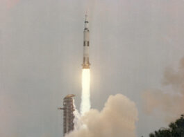 Apollo 13 Blasted Off 55 Years Ago – NASA Anniversary Apollo 13 launches upward, leaving a column of flames and light brown vapor behind it. Trees and vegetation frame the water at the bottom of the photo. Outlines of small animals can be see in the water. At bottom left is written