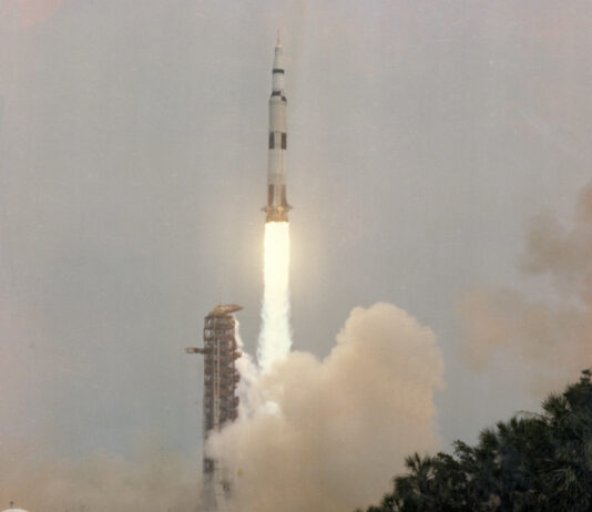 Apollo 13 Blasted Off 55 Years Ago – NASA Anniversary Apollo 13 launches upward, leaving a column of flames and light brown vapor behind it. Trees and vegetation frame the water at the bottom of the photo. Outlines of small animals can be see in the water. At bottom left is written