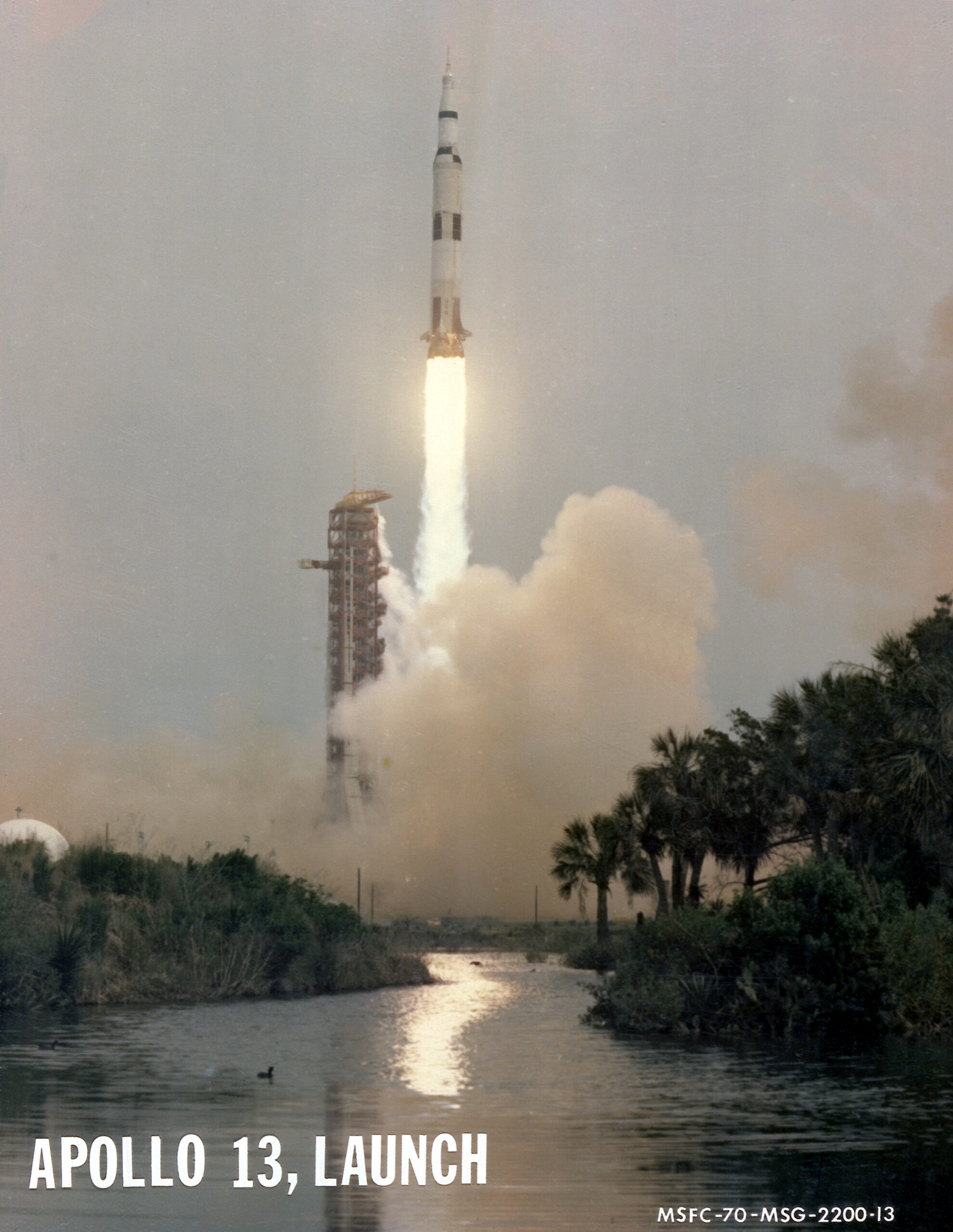 0101247orig 1.jpg Apollo 13 launches upward, leaving a column of flames and light brown vapor behind it. Trees and vegetation frame the water at the bottom of the photo. Outlines of small animals can be see in the water. At bottom left is written