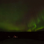 NASA Plans Triple Rocket Launch from Alaska for Aurora Study A time-lapse of auroral activity looks like green, red, yellow, and orange curtains traveling across a clear night sky filled with stars. In the dark land below, faraway launch pads are lit by floodlights, and two tiny vertical rockets can be seen waiting for launch.