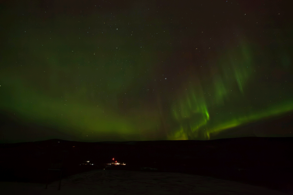 03 aurora.gif A time-lapse of auroral activity looks like green, red, yellow, and orange curtains traveling across a clear night sky filled with stars. In the dark land below, faraway launch pads are lit by floodlights, and two tiny vertical rockets can be seen waiting for launch.