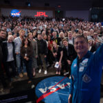 NASA’s Project Hail Mary Film Inspired by Exploration, Science At right, a man in a blue flight suit with patches on it takes a selfie with his cell phone. He is in the photo, as well as panel members and the large audience. They are in an auditorium. In the far background, there is a NASA