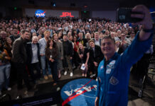 NASA’s Project Hail Mary Film Inspired by Exploration, Science At right, a man in a blue flight suit with patches on it takes a selfie with his cell phone. He is in the photo, as well as panel members and the large audience. They are in an auditorium. In the far background, there is a NASA