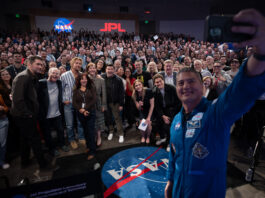NASA’s Project Hail Mary Film Inspired by Exploration, Science At right, a man in a blue flight suit with patches on it takes a selfie with his cell phone. He is in the photo, as well as panel members and the large audience. They are in an auditorium. In the far background, there is a NASA