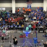 NASA JPL-Backed Robotics Competition Engages Students A robotics competition in a gymnasium with teams in colorful shirts cheering. Robots compete on the field, scoring with blue balls and PVC structures. Referees and volunteers oversee the event.