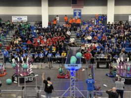 NASA JPL-Backed Robotics Competition Engages Students A robotics competition in a gymnasium with teams in colorful shirts cheering. Robots compete on the field, scoring with blue balls and PVC structures. Referees and volunteers oversee the event.