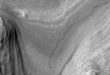 NASA Orbiter Captures Curiosity Rover’s Journey to New Site High-resolution grayscale image of Mars showing layered terrain with smooth slopes, rocky ridges, and a small dark speck near the center, identified as NASA