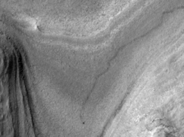 NASA Orbiter Captures Curiosity Rover’s Journey to New Site High-resolution grayscale image of Mars showing layered terrain with smooth slopes, rocky ridges, and a small dark speck near the center, identified as NASA