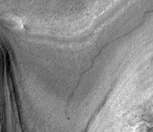 NASA Orbiter Captures Curiosity Rover’s Journey to New Site High-resolution grayscale image of Mars showing layered terrain with smooth slopes, rocky ridges, and a small dark speck near the center, identified as NASA