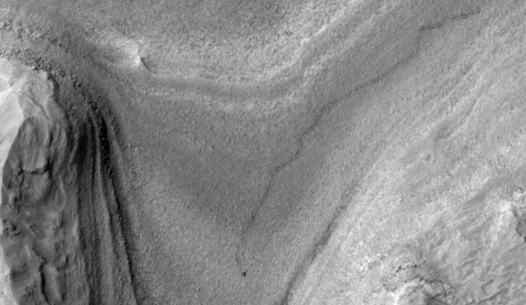 High-resolution grayscale image of Mars showing layered terrain with smooth slopes, rocky ridges, and a small dark speck near the center, identified as NASA