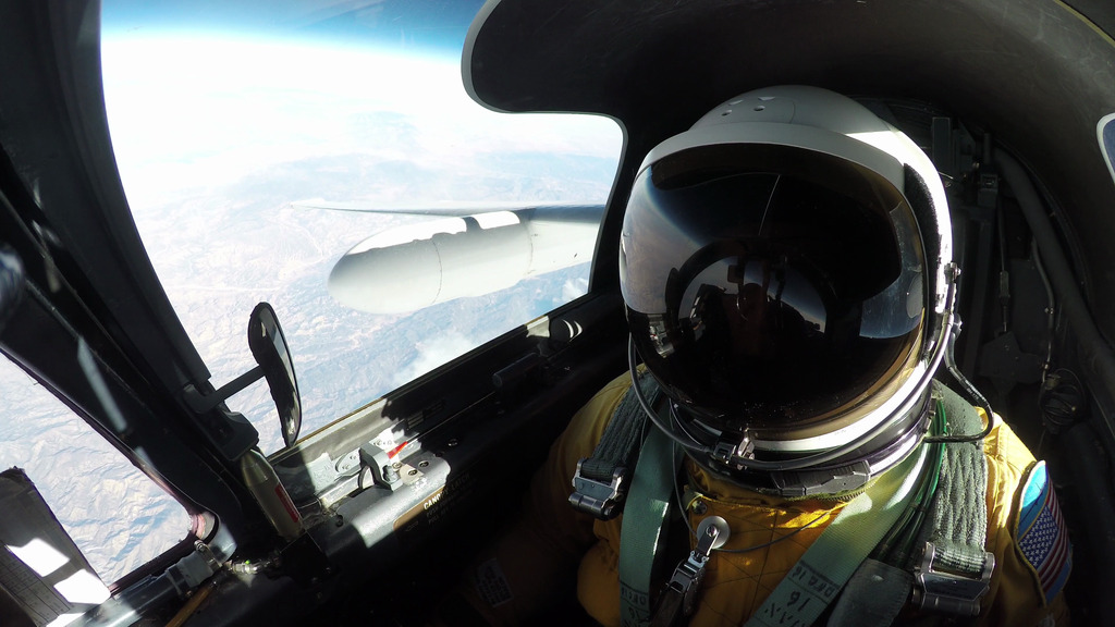 A high-angle, eye-level shot from inside the cockpit of an ER-2 aircraft shows a pilot in a full pressure suit and helmet, with the right wing of the plane and the Earth