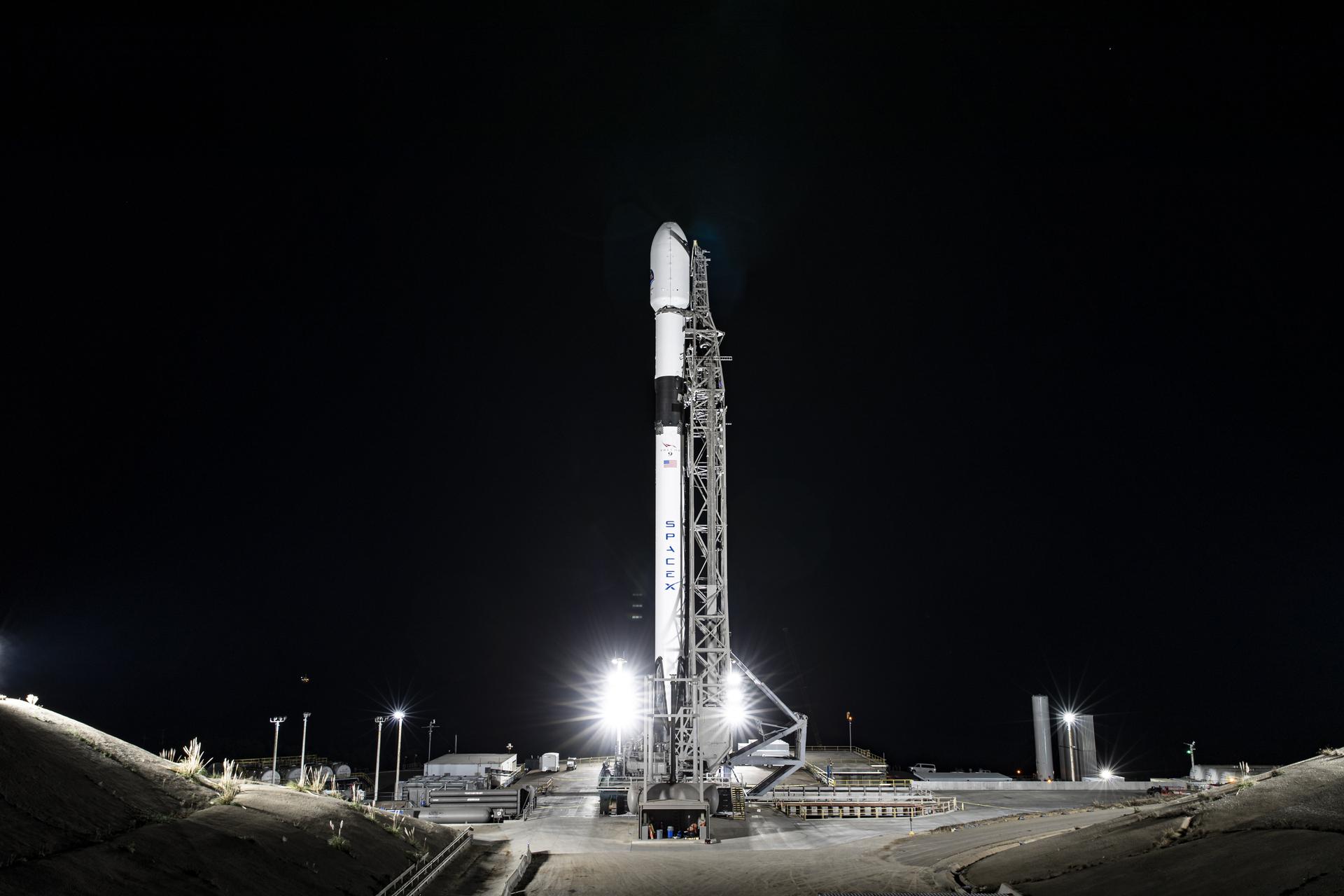 1 s6 mf on pad.jpg A wide-angle, distant photo of a white rocket on the launch pad, brightly lit, with a black night sky behind it.