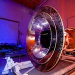 NASA Opens Media Access for Cosmic and Solar Missions Launch The large, shiny silver, round SPHEREx telescope sits in a room lit by blue and orange lighting.