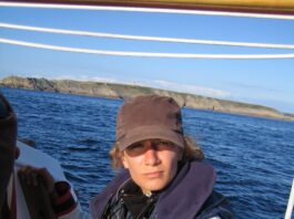 Meloë Kacenelenbogen Focuses on Advancing Air Quality, Climate Studies Meloë S. Kacenelenbogen looks at the camera on a boat with blue water and land in the background. She is wearing a black hat and a red, white, and blue jacket that has a logo with a globe and the words