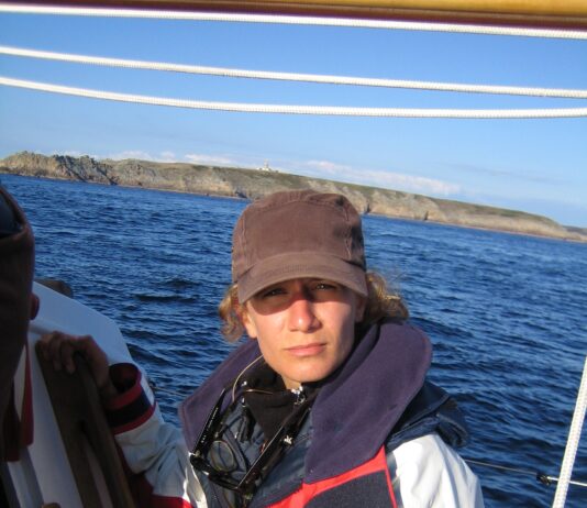 Meloë Kacenelenbogen Focuses on Advancing Air Quality, Climate Studies Meloë S. Kacenelenbogen looks at the camera on a boat with blue water and land in the background. She is wearing a black hat and a red, white, and blue jacket that has a logo with a globe and the words
