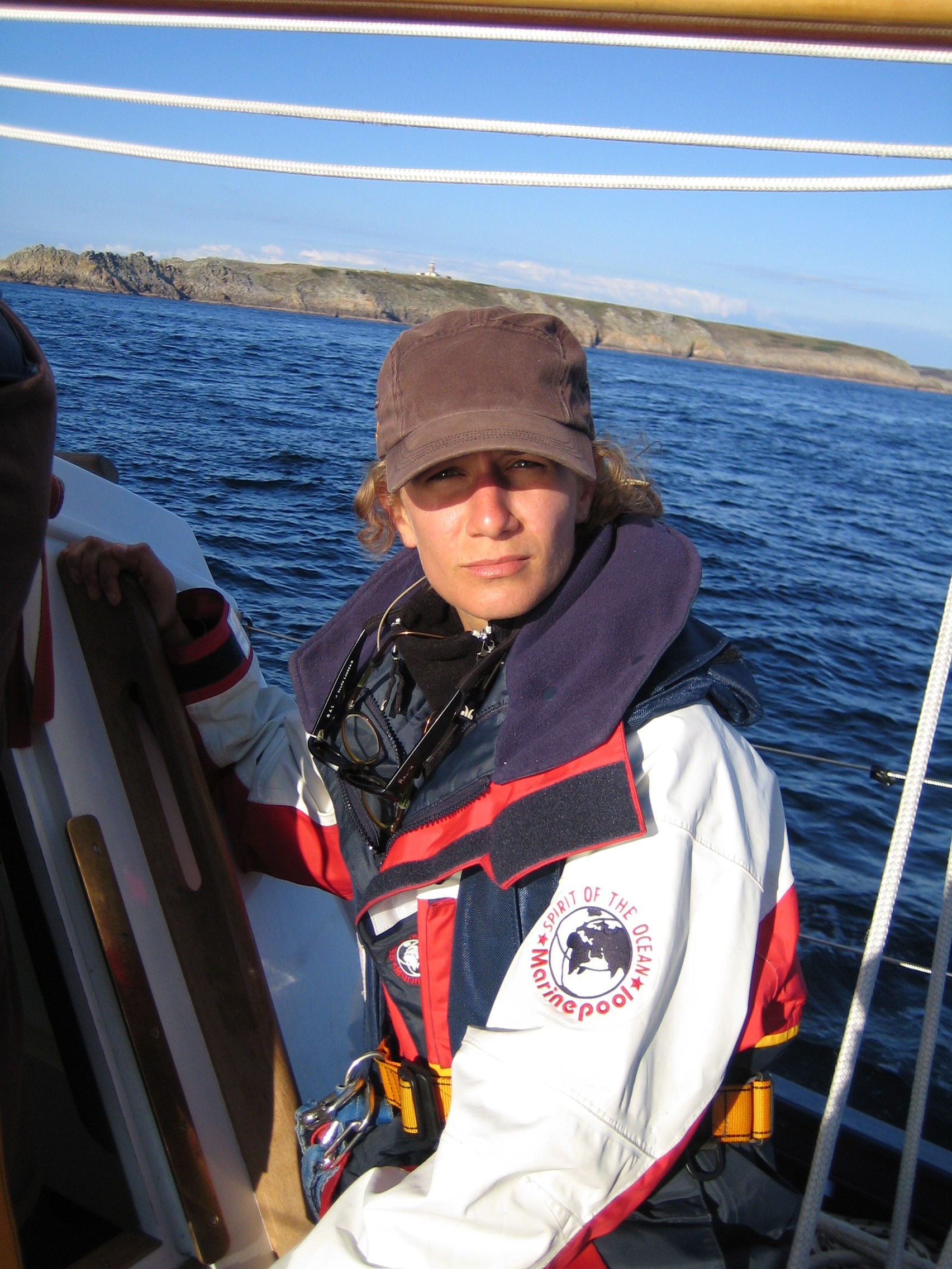 1729616691 3 2.jpg Meloë S. Kacenelenbogen looks at the camera on a boat with blue water and land in the background. She is wearing a black hat and a red, white, and blue jacket that has a logo with a globe and the words