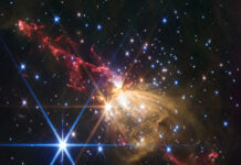 NASA’s Stellar Jet Discovery Gaseous yellow-orange filaments look like a flower seen from the side and tilted slightly from upper left to lower right, slightly higher than the center of the frame. Extending from the flower to upper left and lower right are red gas outflows that have an overall shape of tall, narrow triangles with rounded tips. Each red triangle is made up of wavy, irregular lines. Dozens of stars are scattered across the field. One particularly bright white star with eight diffraction spikes is located at the top of the yellow rose. Another bright blue star with even more prominent diffraction spikes is to its lower left. The background of space is black.