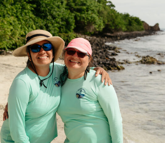 Discussion with NASA’s OCEANOS Expert María Fernanda Barbarena-Arias Two Puerto Rican women stand on a beach, both wearing teal longsleeve shirts and sunglasses. The one on the left has a tan, wide-brimmed hat, and the one on the right has a pink baseball cap.
