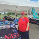 Tina Preyan Drives Innovation at NASA’s Johnson Space Center Johnson Space Center Food Services Specialist Tina Preyan stands in front of a NASA Store table at an outdoor event.