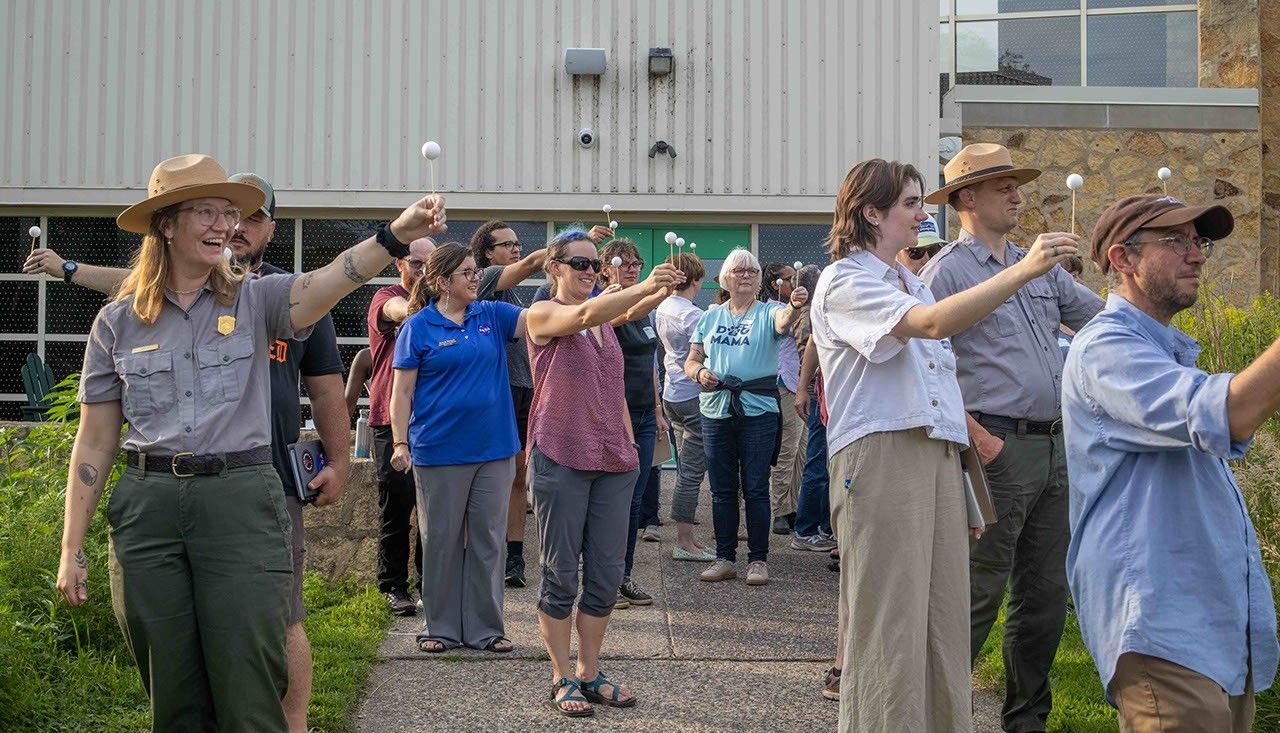 2025 0805 riversofstarsandstories 30 moonactivity sm brandi stewart.jpg A group of people stand outside holding white Styrofoam balls on a stick with arms outstretched to model moon phases.