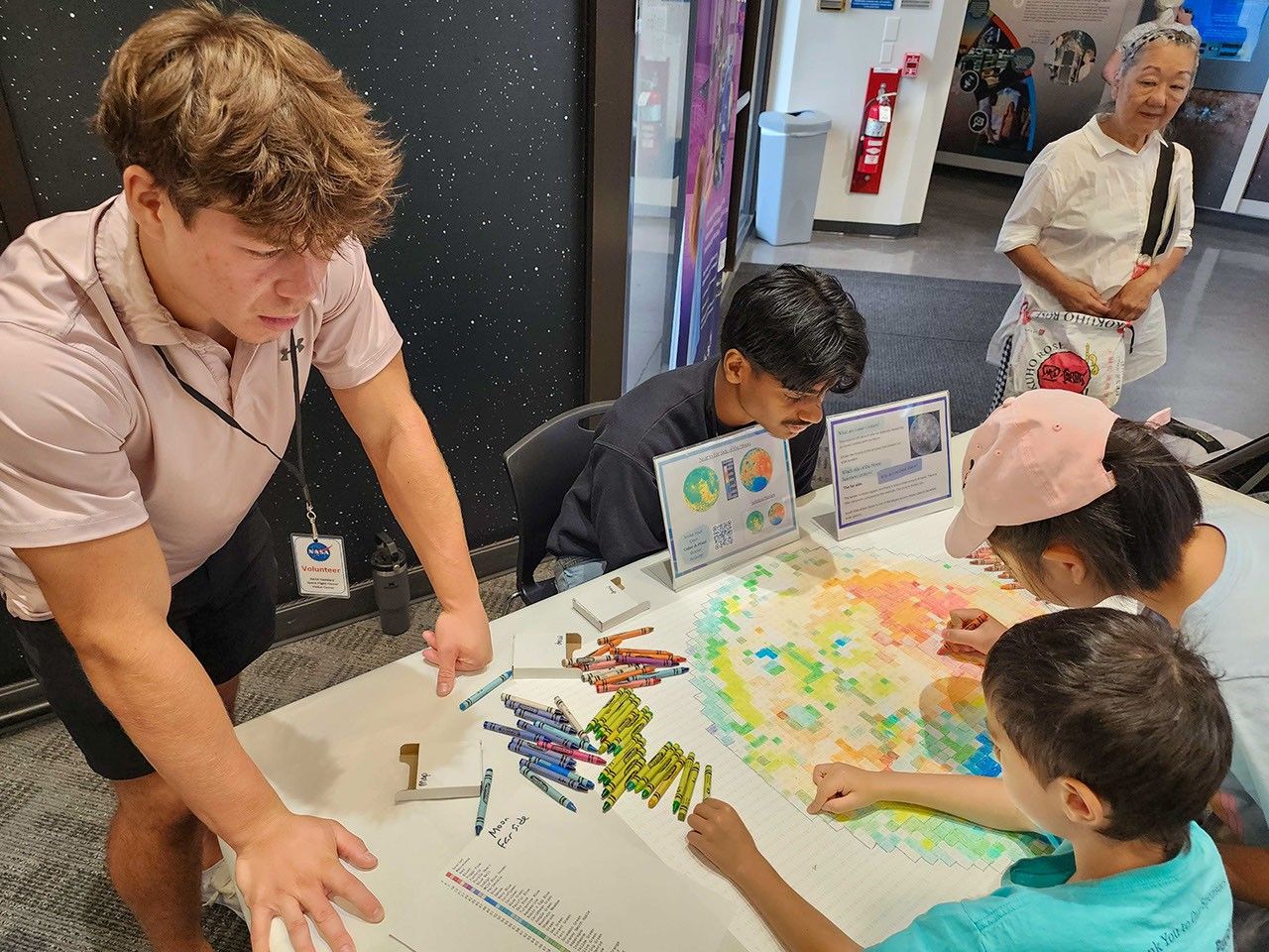 People gather around a pixelated poster of the Moon’s far side and color in the small boxes with a crayon color that corresponds to the elevation in that area.