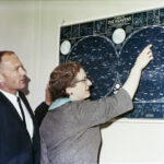 NASA Celebrates 100 Years Since Nancy Grace Roman’s Birth Two people - astronaut Buzz Aldrin and Dr. Nancy Grace Roman - look at a map of celestial objects hanging on a wall in front of them. The photos is taken from the side, so their profiles are visible. Aldrin wears a dark suit and tie, while Roman wears a gray short sleeved jacket. She is pointing at a constellation on the blue and white map.