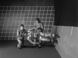 NASA Breaks Barriers with Anti-Gravity Innovations In this black and white photo, two people adjust a 1960s-era spacesuit on a person who is suspended by wires. This third person is parallel to the floor, with his feet on the wall on the right.