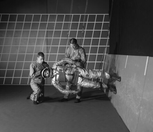 NASA Breaks Barriers with Anti-Gravity Innovations In this black and white photo, two people adjust a 1960s-era spacesuit on a person who is suspended by wires. This third person is parallel to the floor, with his feet on the wall on the right.