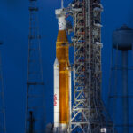 NASA Calls Media to Artemis II Lunar Mission Launch A large orange NASA rocket with white booster rockets stands vertically on a launch pad, lit by spotlights, with the full Moon and night sky behind it.