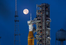 NASA Calls Media to Artemis II Lunar Mission Launch A large orange NASA rocket with white booster rockets stands vertically on a launch pad, lit by spotlights, with the full Moon and night sky behind it.