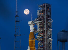 NASA Calls Media to Artemis II Lunar Mission Launch A large orange NASA rocket with white booster rockets stands vertically on a launch pad, lit by spotlights, with the full Moon and night sky behind it.