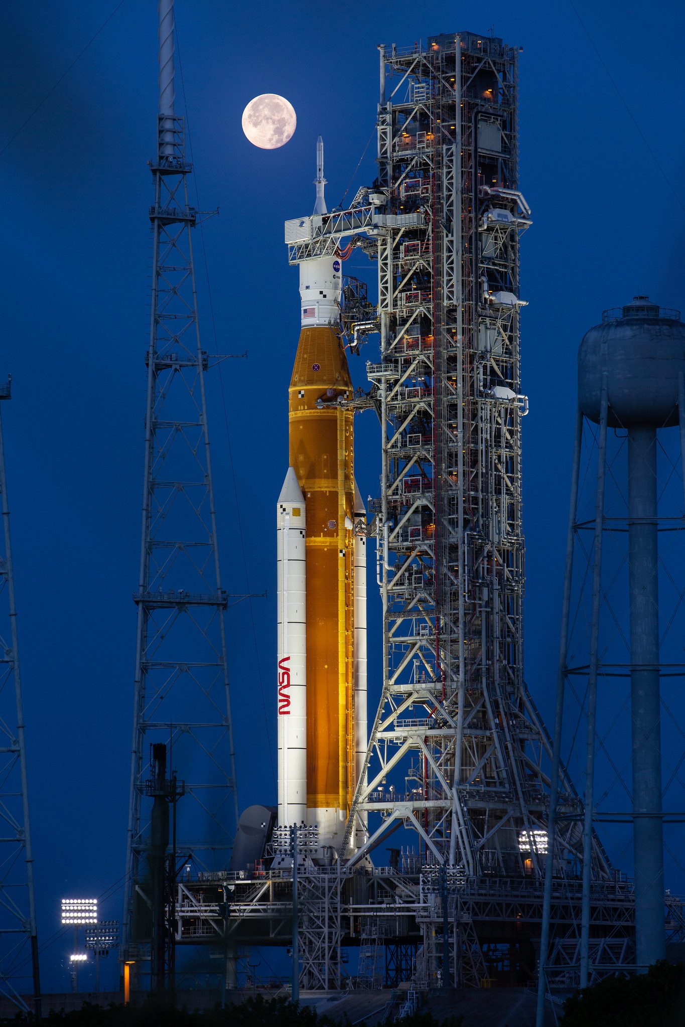 52148647216 95f46d2378 k.jpg A large orange NASA rocket with white booster rockets stands vertically on a launch pad, lit by spotlights, with the full Moon and night sky behind it.