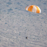 NASA’s OSIRIS-REx Mission Returns for New Exploration Insights An orange and white parachute is visible at top right. It is attached to a training model of a capsule. The black and white cone-like capsule is quite small in this image. The pale ground below makes up the background of the image.