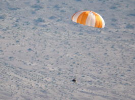 NASA’s OSIRIS-REx Mission Returns for New Exploration Insights An orange and white parachute is visible at top right. It is attached to a training model of a capsule. The black and white cone-like capsule is quite small in this image. The pale ground below makes up the background of the image.