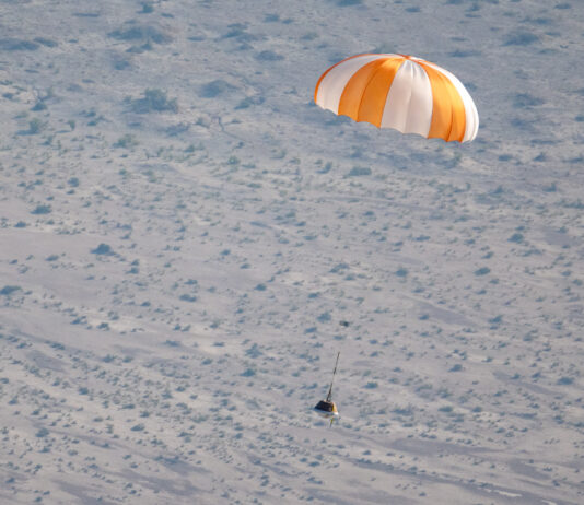 NASA’s OSIRIS-REx Mission Returns for New Exploration Insights An orange and white parachute is visible at top right. It is attached to a training model of a capsule. The black and white cone-like capsule is quite small in this image. The pale ground below makes up the background of the image.