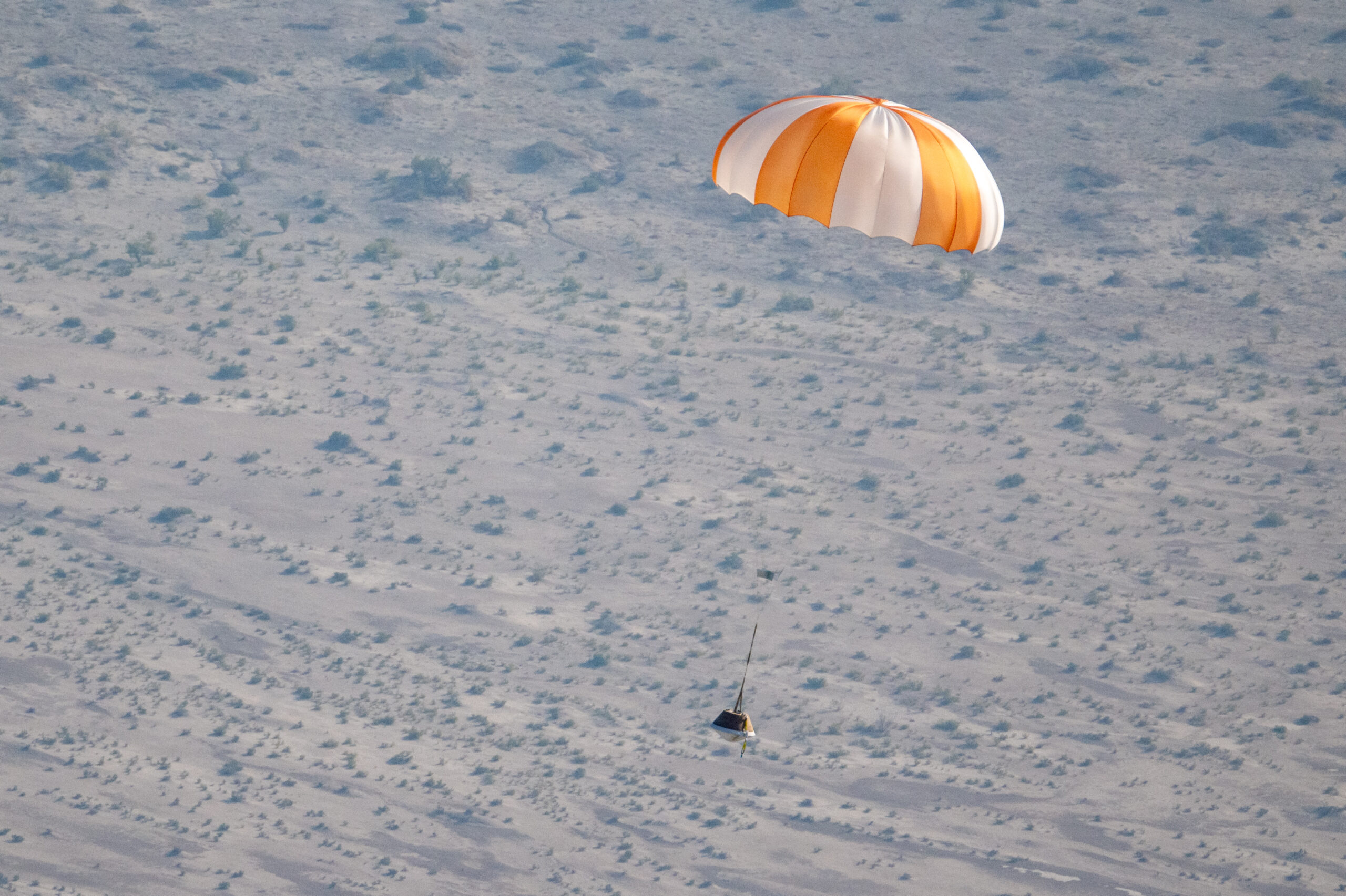 53153681225 92ea082697 o.jpg An orange and white parachute is visible at top right. It is attached to a training model of a capsule. The black and white cone-like capsule is quite small in this image. The pale ground below makes up the background of the image.