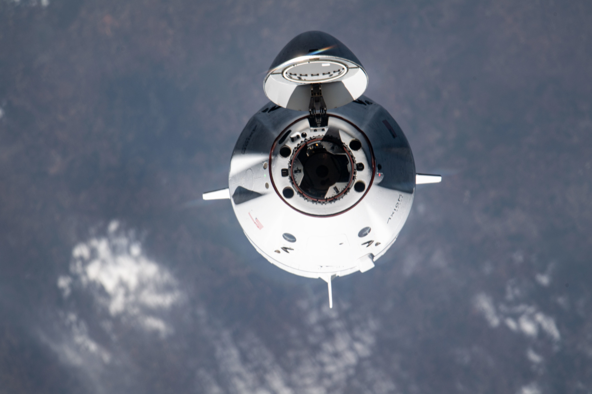 A white spacecraft faces the camera with its nose cone open, with Earth in the background.