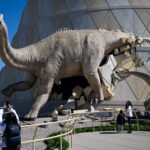 NASA’s Dinosaur Ready for Safe Solar Eclipse Viewing 2024 A large model dinosaur bursts out of a beige and gray building. The Alamosaurus wears a pair of eclipse glasses. In the foreground, a child lifts their phone to take a picture of the dinosaur.