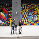 NASA Revives Art Program with NYC Mural in 2024 Two adults and a child pose for a photo in front of a colorful space-themed mural. The child wears an astronaut costume.