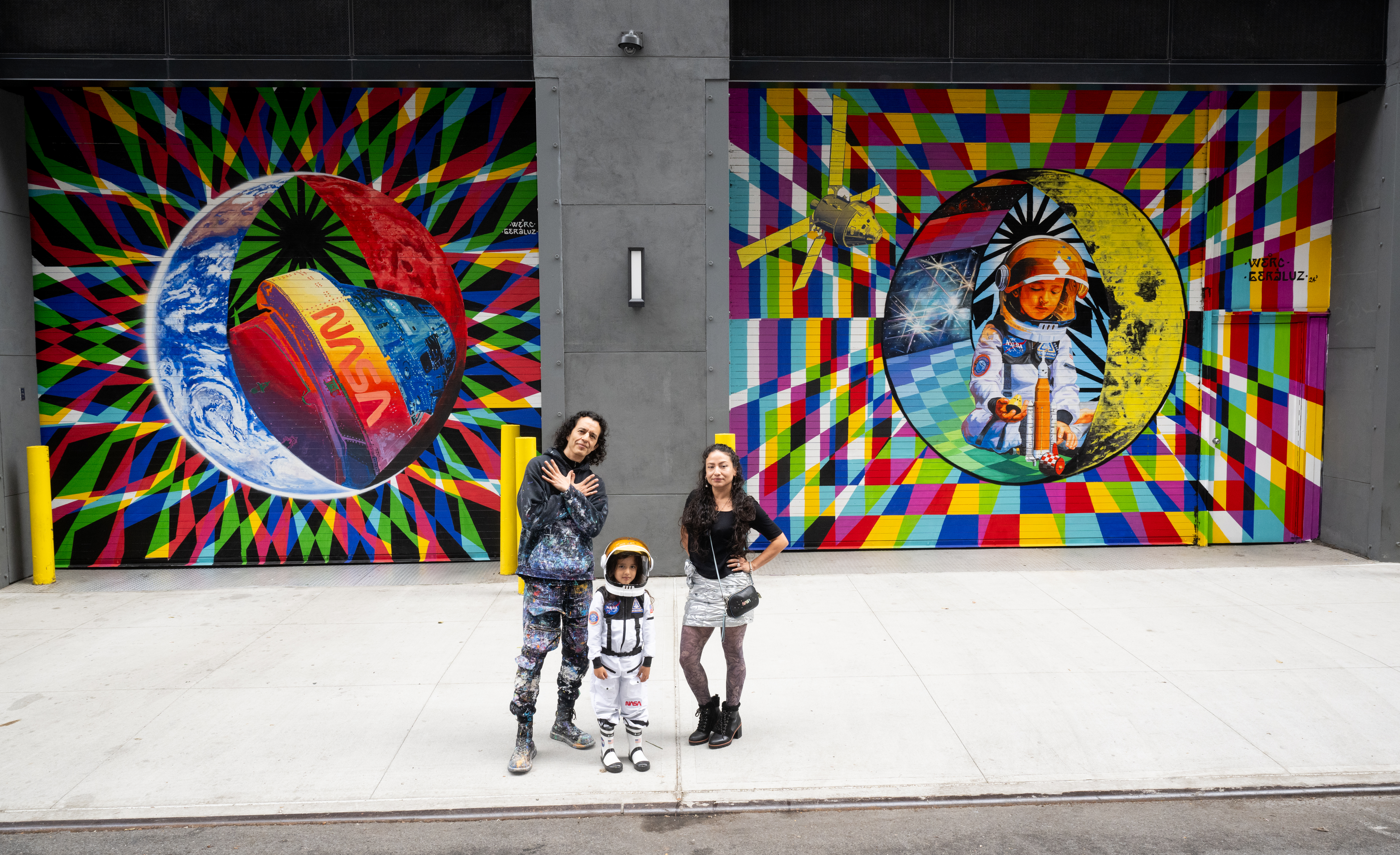 54023707668 041ea3961a o.jpg Two adults and a child pose for a photo in front of a colorful space-themed mural. The child wears an astronaut costume.