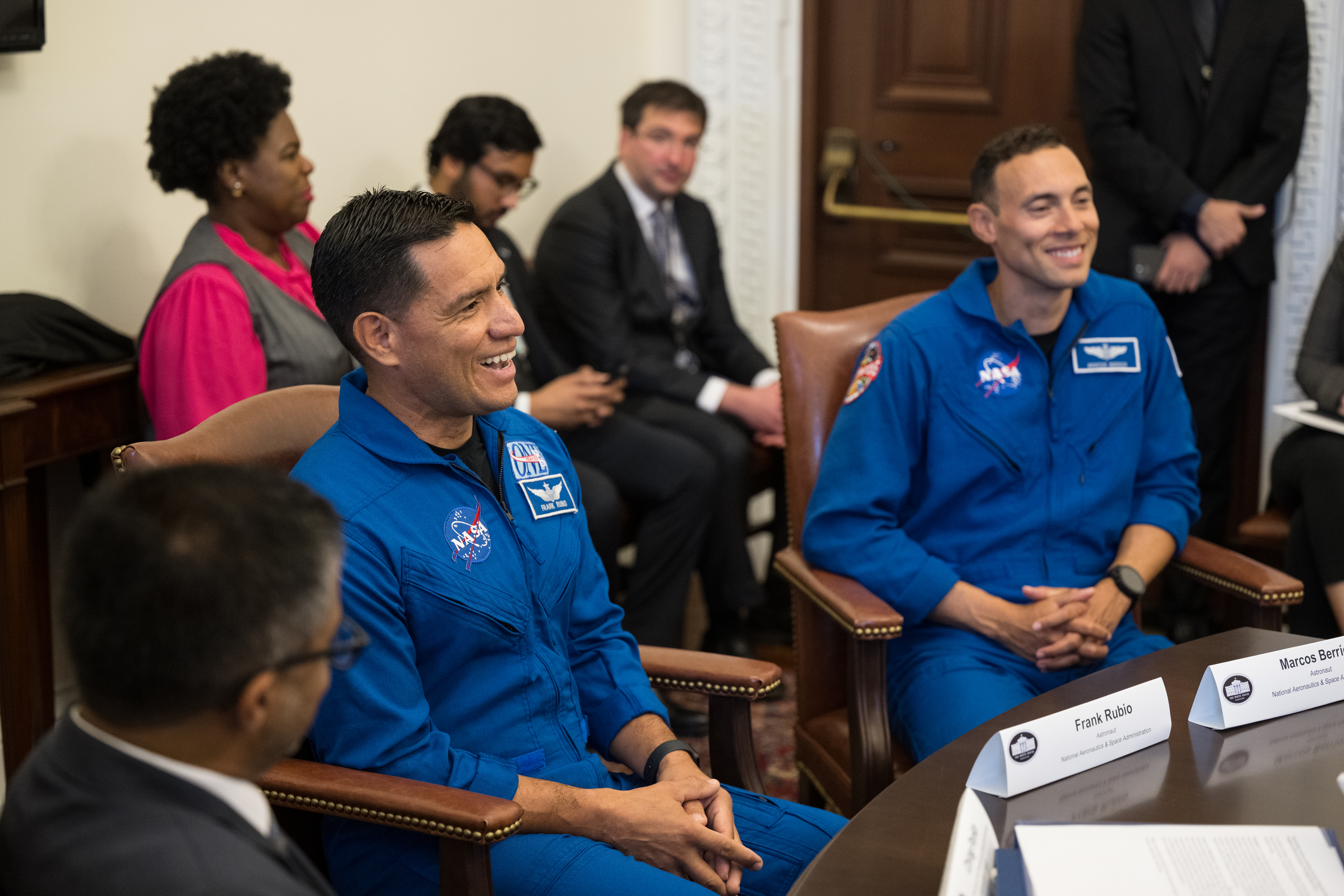 54033691286 310028c34f o.jpg Two Hispanic men in blue jumpsuits with NASA patches on them sit at a table, smiling at people off screen. There are people of different ethnicities in the background.