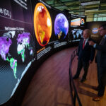 Smithsonian Hosts NASA’s Earth Information Center Exhibit Two men in suits stand behind a short curved barrier. They look at a large curved screen that spans the room. The screen shows information, maps, and other graphics that relay information about carbon dioxide. The man on the right is pointing at the display.