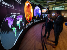 Smithsonian Hosts NASA’s Earth Information Center Exhibit Two men in suits stand behind a short curved barrier. They look at a large curved screen that spans the room. The screen shows information, maps, and other graphics that relay information about carbon dioxide. The man on the right is pointing at the display.