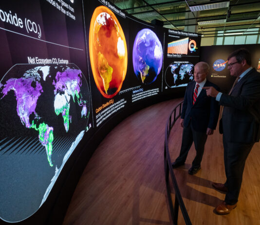 Smithsonian Hosts NASA’s Earth Information Center Exhibit Two men in suits stand behind a short curved barrier. They look at a large curved screen that spans the room. The screen shows information, maps, and other graphics that relay information about carbon dioxide. The man on the right is pointing at the display.