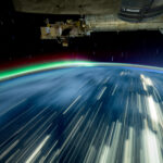 NASA’s 2024 Milestones on the International Space Station Streaks of white light cover a blue Earth, and a green and red aurora rises from the horizon. The exterior of the space station is visible in the top third of the image.