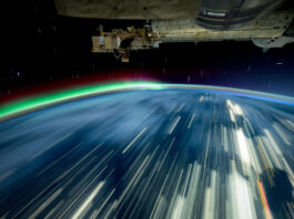 NASA’s 2024 Milestones on the International Space Station Streaks of white light cover a blue Earth, and a green and red aurora rises from the horizon. The exterior of the space station is visible in the top third of the image.