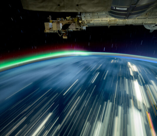 NASA’s 2024 Milestones on the International Space Station Streaks of white light cover a blue Earth, and a green and red aurora rises from the horizon. The exterior of the space station is visible in the top third of the image.