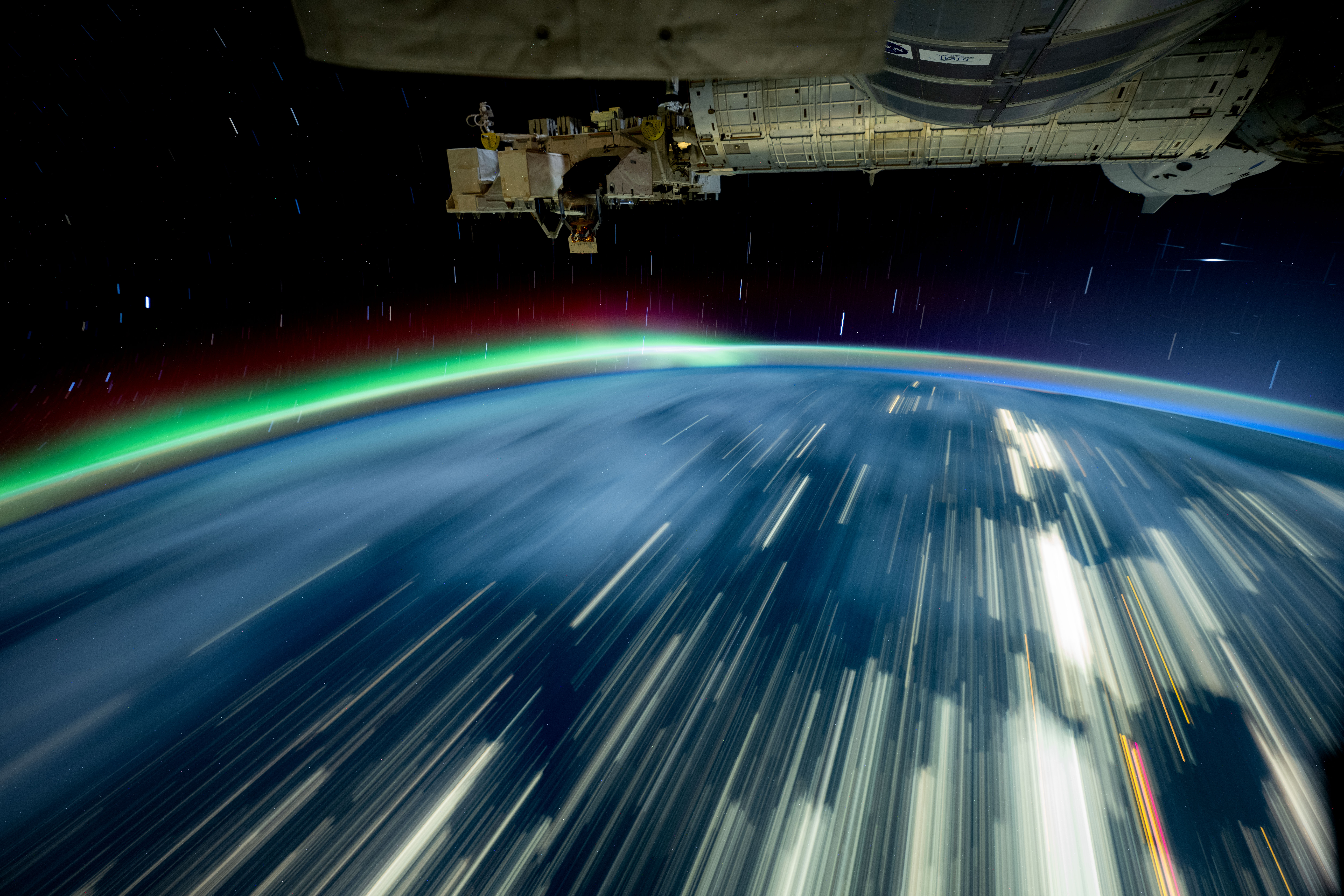 54099828916 45c9bee307 o.jpg Streaks of white light cover a blue Earth, and a green and red aurora rises from the horizon. The exterior of the space station is visible in the top third of the image.