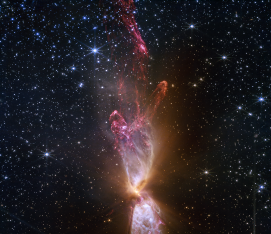 NASA: As Time Passes, Discoveries Continue At the center is a thin vertical cloud known as Lynds 483 that is shaped like an hourglass with irregular edges. At lower center are two discrete bright white, tiny blobs of light that have raced away from the hidden central stars. The top lobe shows a more prominent orange U-shape. Orange bleeds into light purple, and brighter pink at its edges. Some background stars are visible through sections of this lobe. Higher up, there is an orange arc. Some brighter pink material extends to the top edges near the center. In the lower lobe, less orange is visible. More opaque light purple is in its top third, rippling out into semi-transparent blues and pinks. The lower lobe has more texture. V-shapes left and right of the lobes are darkest, and the background stars in these areas appear orange. Elsewhere, the black background of space is clearer, speckled with tiny white stars and faint orange galaxies.